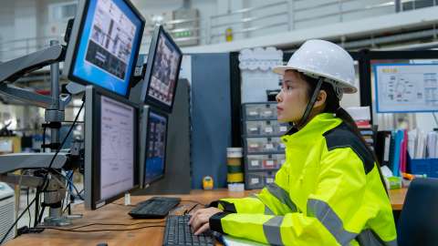 An employee in a manufacturing plant, wearing a hard hat and safety yellow jacket, sits at a desk reviewing manufacturing workflows and graphs shown on four mounted display monitors