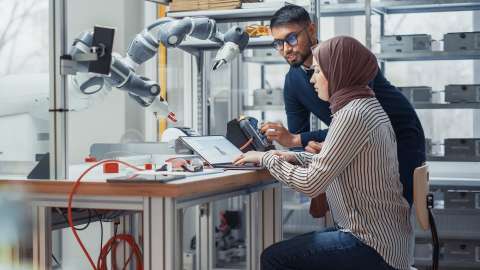 Two students work in an engineering lab. One student sits at a table using a laptop to modify a 3D model, while the other student stands and uses a physical control box to operate robotic arms mounted to the wall above the desk